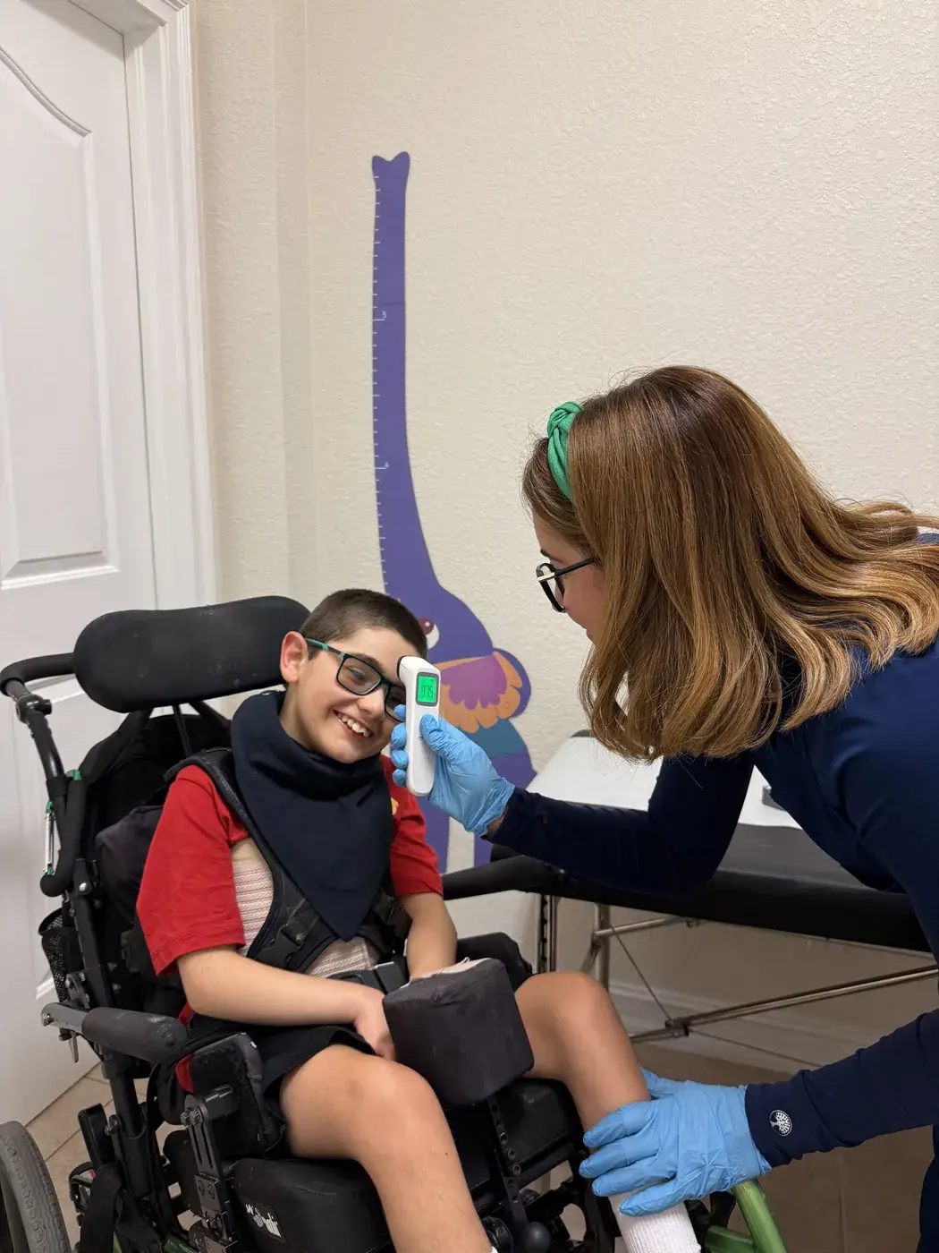 A nurse taking the temperature of a smiling boy in a wheelchair in the nursing area at Ellie's World PPEC in Port Charlotte, Florida