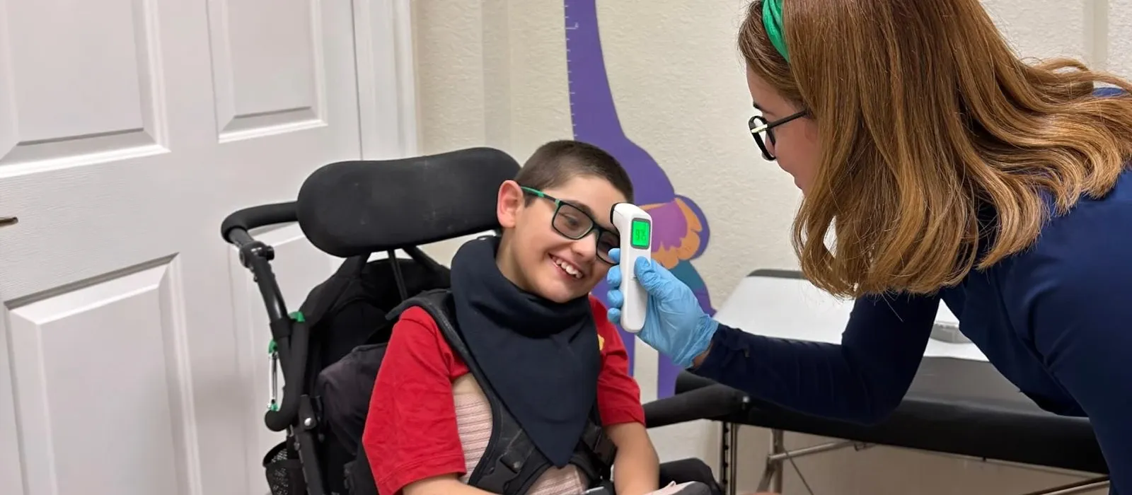A licensed nurse taking the temperature of a smiling boy in a wheelchair at Ellie's World PPEC, a pediatric medical daycare in Port Charlotte, Florida