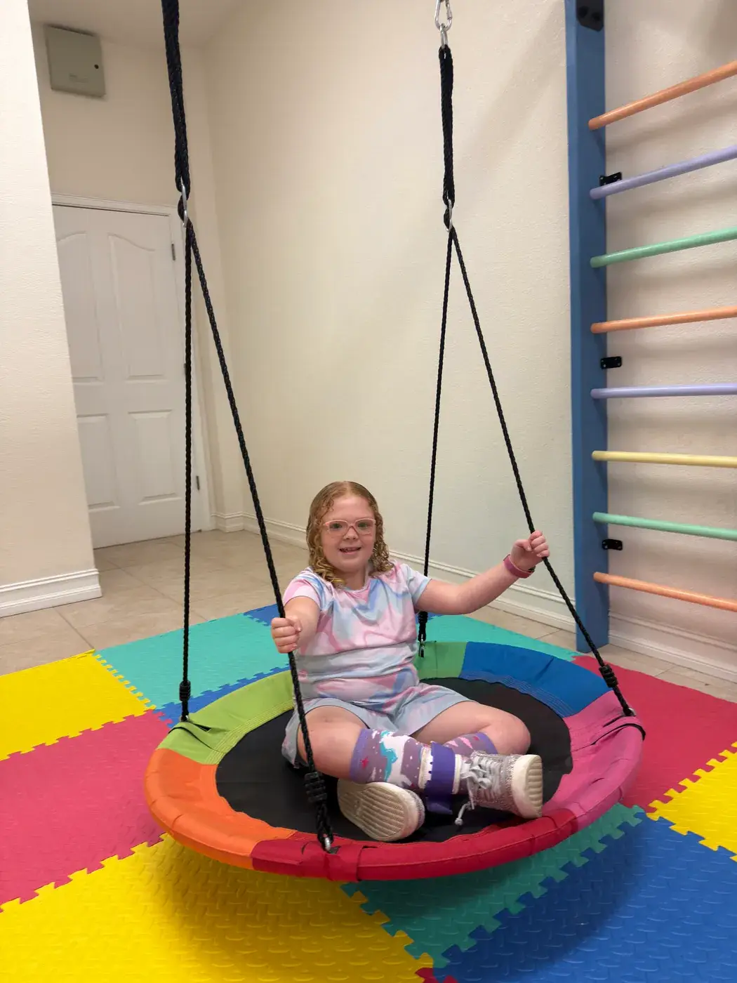 A girl smiling on the indoor platform swing in the sensory room at Ellie's World PPEC in Port Charlotte, Florida